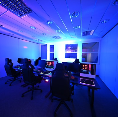 A modern gaming setup with multiple high-backed gaming chairs and monitors arranged in a room with a cool blue lighting. The computers have illuminated keyboards and glowing screens, creating a high-tech ambiance.
