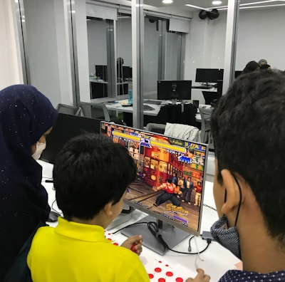A group of children is gathered around a desktop computer, engaged in playing a colorful, action-packed arcade game displayed on the monitor. The setting appears to be a modern, well-lit office or gaming space with large glass windows, showing multiple computer workstations in the background.