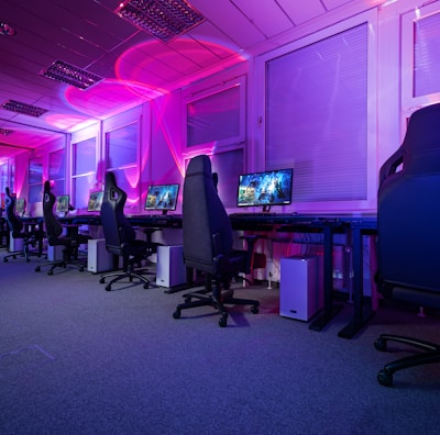A row of gaming computers with large monitors and racing-style chairs is set up along the interior of a modern room. The space is illuminated with vibrant purple and blue LED lighting, creating a futuristic atmosphere. The room is organized and clean, suggesting a professional gaming or esports environment.