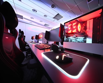 A row of high-end gaming computers on sleek desks, featuring RGB lighting and gaming accessories. The computers are housed in transparent cases with visible components, emitting a red glow. Comfortable gaming chairs with ergonomic design are positioned next to the desks, and the room's ceiling is modern with recessed lighting.