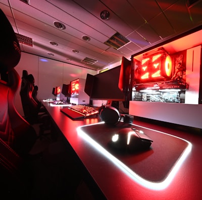 A modern gaming setup featuring several high-end computers with illuminated red interior lighting. The room is equipped with gaming chairs exhibiting a sleek black and red design. Multiple monitors are arranged on a long desk, and peripheral devices like keyboards and mice are uniformly placed, reflecting an ambiance ideal for gaming or competitive play.