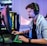 A young person is intensely focused on a computer screen while wearing a headset with a microphone. They are using a gaming keyboard and mouse, sitting in a gaming chair. The room has cool lighting, with a visible PC tower.