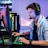A young person is intensely focused on a computer screen while wearing a headset with a microphone. They are using a gaming keyboard and mouse, sitting in a gaming chair. The room has cool lighting, with a visible PC tower.