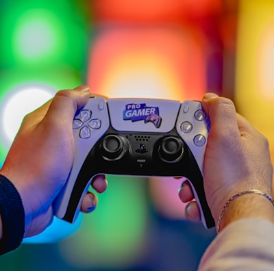 Two hands are holding a gaming controller labeled 'Pro Gamer'. The background is blurred with colorful lights creating a vibrant atmosphere.